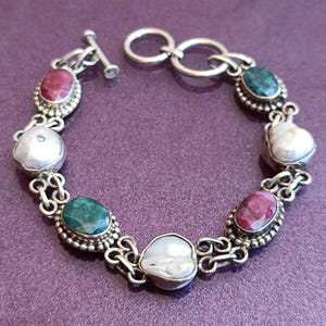 May include: A silver bracelet with a toggle clasp features alternating oval-shaped green, pink, and white gemstones.