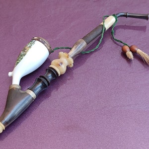 May include: A vintage porcelain pipe with a wooden stem and a green cord with brown tassels. The pipe has a white porcelain bowl with a floral design and a brown wooden mouthpiece.