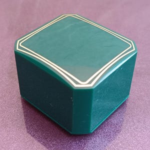 Puede incluir: Una caja verde oscuro en forma de diamante con un borde dorado. La caja tiene una base cuadrada y una tapa a juego. La caja es probablemente para joyas u otros objetos peque&ntilde;os.
