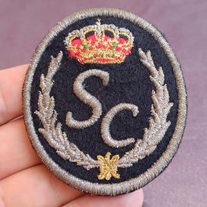 Op de afbeelding: Een ronde, zwart geborduurde patch met de zilveren letters SC in het midden. Boven de letters staat een rood-gouden kroon en eronder een kleine gouden ster. Het ontwerp is omlijst door zilveren lauriertakken en een zilveren randafwerking.