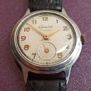 Puede incluir: Un reloj de pulsera plateado con una esfera blanca y una correa de cuero negra. La esfera del reloj tiene números y manecillas de color dorado. La esfera del reloj también tiene el texto "Слава 17 КАМНЕЙ" y "ЗАВОД НАС 3А".