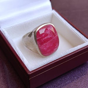 Op de afbeelding: Een zilveren ring met een grote, rechthoekige, gefacetteerde, roze edelsteen. De ring wordt gepresenteerd in een rode sieradendoos met een wit interieur. De edelsteen heeft een gemarmerd uiterlijk.