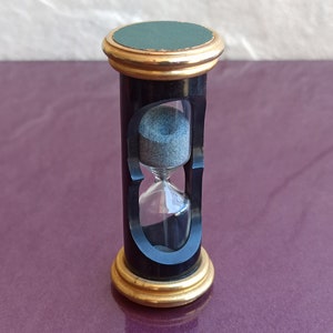 以下が含まれることがあります： 金色の縁取りと緑色の天蓋が付いた黒い砂時計。砂時計は砂で満たされ、紫色の表面の上に置かれています。