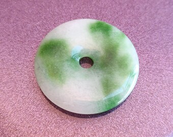 May include: A white jade pendant with green swirls. The pendant is shaped like a donut with a hole in the center.