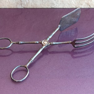 Puede incluir: Pinzas de servir plateadas con un mango de estilo bambú y una cabeza plana de forma ovalada. Las pinzas tienen un diseño de tenedor en un extremo.