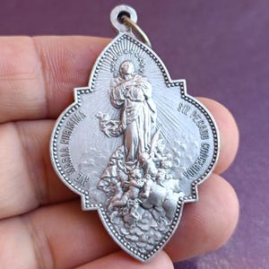 May include: Silver-toned religious pendant with a detailed depiction of a saint, surrounded by text. The pendant has a decorative border and a small loop at the top for attachment.