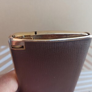 May include: A brown leather cigarette case with a gold metal trim. The case has a textured surface and a small knob on the side.