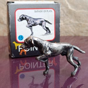 Può includere: Figurina in argento di un cane che punta in una posa di caccia. La figurina è in piedi su una superficie viola. La scatola in cui era contenuta è visibile sullo sfondo. La scatola è nera con testo bianco che dice "Bañado en Plata" e "15".