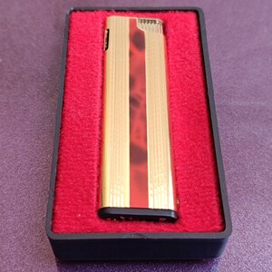 Peut inclure: Un briquet doré avec une bande verticale rouge et marron, présenté dans un étui noir avec un intérieur en velours rouge. Le briquet est de forme rectangulaire et a une surface texturée.