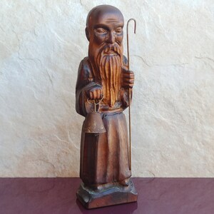 May include: A wooden figurine of a bearded man holding a bell and a staff. The figurine is carved in a dark brown wood and has a detailed face.