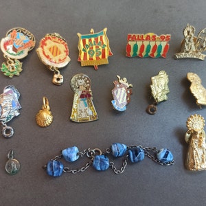 May include: A collection of 15 vintage enamel pins and charms, including religious figures, a seashell, a crown, and a Spanish flag. The pins are colourful and feature various designs.
