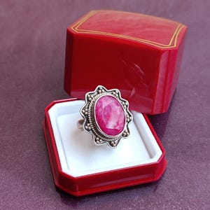 Op de afbeelding: Een zilveren ring met een grote, ovale, roze edelsteen wordt getoond in een rode sieradendoos. De edelsteen is omgeven door een decoratieve zilveren zetting met een kralenrand. De sieradendoos is open en onthult de ring tegen een witte achtergrond.