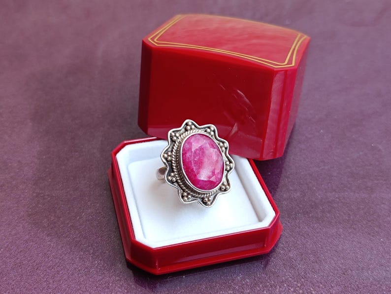 Op de afbeelding: Een zilveren ring met een grote, ovale, roze edelsteen wordt getoond in een rode sieradendoos. De edelsteen is omgeven door een decoratieve zilveren zetting met een kralenrand. De sieradendoos is open en onthult de ring tegen een witte achtergrond.
