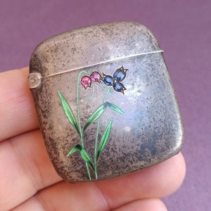 May include: A silver matchbox with a floral design. The flowers are painted in shades of pink, blue, and green. The matchbox has a tarnished finish.