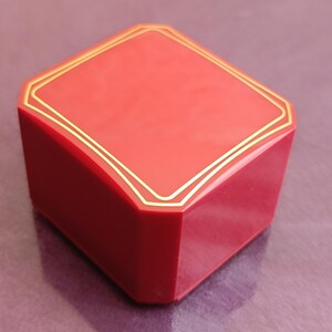 Op de afbeelding: Een rode, ruitvormige doos met een goudkleurige rand rond de bovenkant. De doos heeft een glanzende afwerking en een geometrisch ontwerp. De doos wordt waarschijnlijk gebruikt voor sieraden of kleine items.