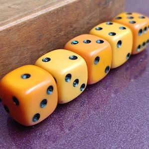 May include: Five vintage dice in a row, each with black dots indicating numbers. The dice are a gradient of orange and yellow, with a wooden block in the background. The dice are a classic game accessory.