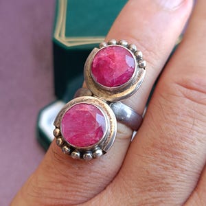 Puede incluir: Anillo de plata con dos grandes piedras preciosas rosas brillantes, de forma ovalada y facetadas. Cada gema est&aacute; engastada en un bisel de plata y rodeada de peque&ntilde;as cuentas de plata. El anillo se muestra en un dedo.