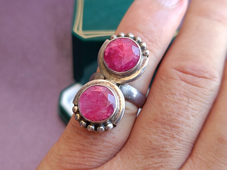 Puede incluir: Anillo de plata con dos grandes piedras preciosas rosas brillantes, de forma ovalada y facetadas. Cada gema est&aacute; engastada en un bisel de plata y rodeada de peque&ntilde;as cuentas de plata. El anillo se muestra en un dedo.