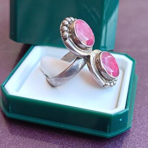 Puede incluir: Un anillo de plata con dos piedras preciosas ovaladas de color rosa brillante. Las piedras est&aacute;n engastadas en un bisel con un borde de cuentas. El anillo se exhibe en una caja de joyer&iacute;a verde y blanca.