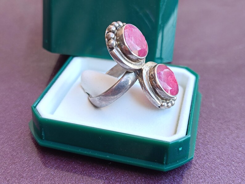 Puede incluir: Un anillo de plata con dos piedras preciosas ovaladas de color rosa brillante. Las piedras est&aacute;n engastadas en un bisel con un borde de cuentas. El anillo se exhibe en una caja de joyer&iacute;a verde y blanca.