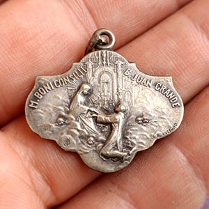 Peut inclure: Un pendentif religieux argenté avec un dessus en forme d'arche et une scène en relief détaillée. La scène représente deux personnages, dont un à genoux, avec du texte autour des bords, dont "M. BONI CONSILII" et "B. JUAN GRANDE". Le pendentif a une boucle pour l'accrocher.