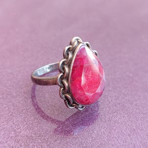 Pode incluir: Um anel de prata com uma gema rosa em forma de l&aacute;grima engastada em uma moldura de prata decorativa.