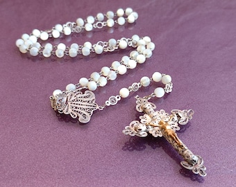 Può includere: Un rosario d'argento con perline bianche di madreperla e una croce in filigrana.