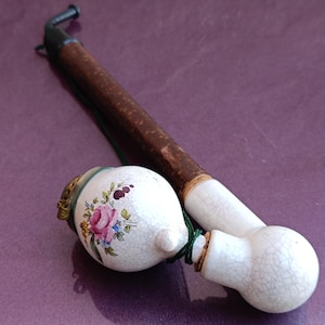 May include: An antique tobacco pipe with a white ceramic bowl decorated with a pink rose and blue floral design. The pipe has a brown wooden stem and a black metal mouthpiece. The pipe is resting on a purple surface.