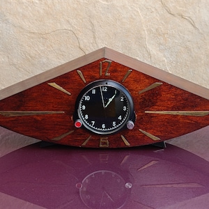 Può includere: Un orologio in legno scuro con un quadrante nero e dettagli dorati. L'orologio ha la forma di un triangolo con bordi arrotondati. Il quadrante dell'orologio ha uno sfondo nero con numeri e lancette bianchi. L'orologio è su una superficie viola.