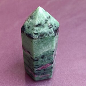 May include: A green and black zoisite crystal point with a faceted top. The crystal has a polished surface and displays streaks of pink and black throughout its structure. The crystal is approximately 3 inches tall.