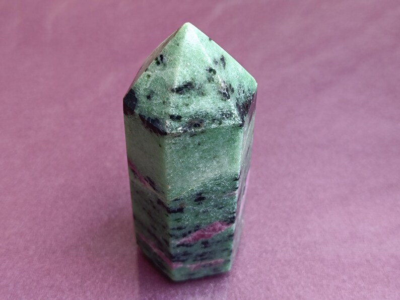 May include: A green and black zoisite crystal point with a faceted top. The crystal has a polished surface and displays streaks of pink and black throughout its structure. The crystal is approximately 3 inches tall.