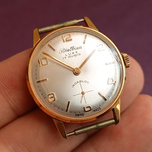 Könnte beinhalten: Eine goldfarbene Armbanduhr mit weißem Zifferblatt und schwarzen Zeigern. Das Zifferblatt der Uhr hat schwarze römische Ziffern als Stundenmarkierungen und einen schwarzen Sekundenzeiger. Das Zifferblatt der Uhr trägt außerdem den Text "Blatina Luxe 17 Rubis Incabloc Swiss Made".