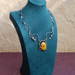 May include: A silver necklace with a large amber stone pendant. The necklace is designed with delicate, flowing lines that resemble leaves.