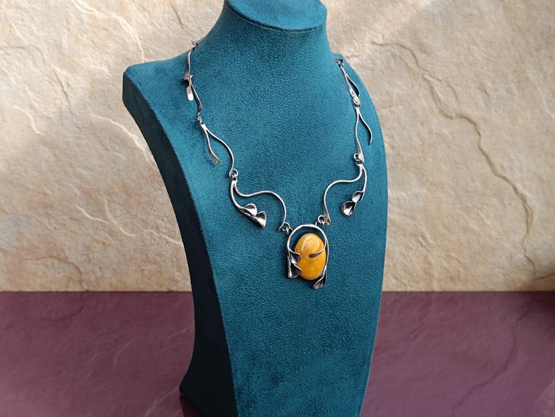 May include: A silver necklace with a large amber stone pendant. The necklace is designed with delicate, flowing lines that resemble leaves.