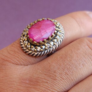 Op de afbeelding: Een zilveren ring met een grote roze ovale edelsteen omringd door kleinere heldere edelstenen. De ring is gezet in een goudkleurig metaal.
