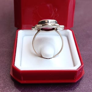 Op de afbeelding: Een zilveren ring met een grote, ovale, roze edelsteen gezet in een bloemmotief. De ring wordt gepresenteerd in een rode sieradendoos met een witte binnenkant, wat een opvallend contrast cre&euml;ert. De ringband is dun en delicaat.