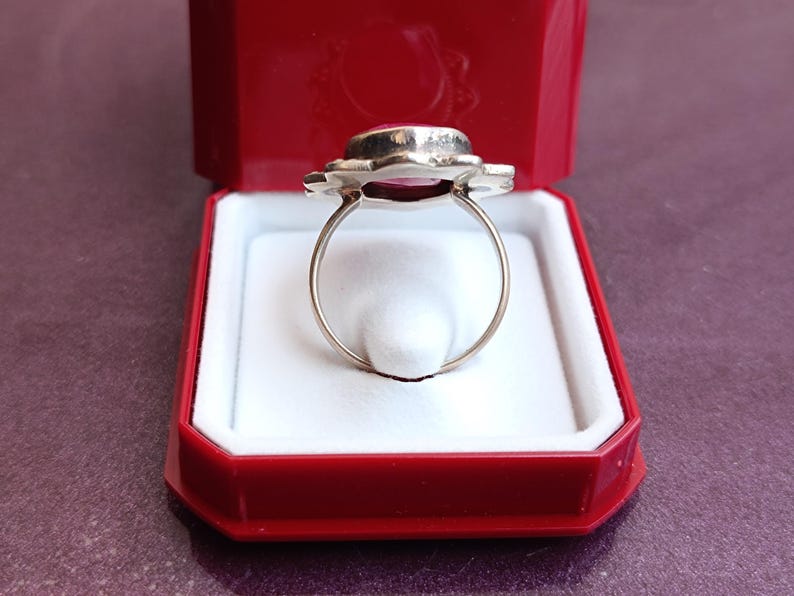 Op de afbeelding: Een zilveren ring met een grote, ovale, roze edelsteen gezet in een bloemmotief. De ring wordt gepresenteerd in een rode sieradendoos met een witte binnenkant, wat een opvallend contrast cre&euml;ert. De ringband is dun en delicaat.
