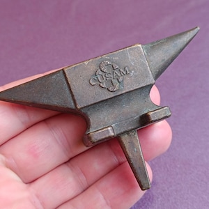 May include: A small, bronze-colored anvil with a pointed horn and a rectangular face. The anvil has a rectangular base with a single, downward-pointing horn. The text "CUSAM" is visible on the face of the anvil.