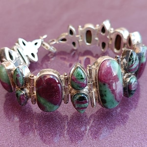 Puede incluir: Pulsera plateada con piedras ovaladas y en forma de lágrima en tonos rojo intenso y verde. La pulsera tiene un cierre decorativo y está sobre un fondo morado.