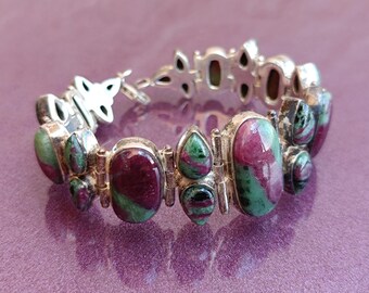 May include: Silver-toned bracelet featuring oval and teardrop-shaped stones with a mix of deep red and green hues. The bracelet has a decorative clasp and is set against a purple background.