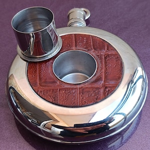Peut inclure: Une flasque à hanche en acier inoxydable poli avec une incrustation circulaire en cuir brun rougeâtre texturé. Une tasse en acier inoxydable assortie est posée sur la flasque, et une petite ouverture ronde est au centre.