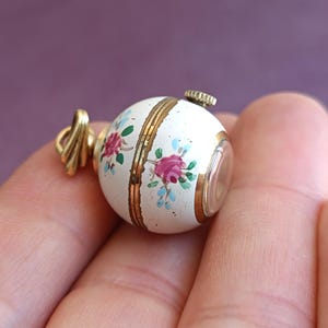 May include: A small, round, white pendant with a floral design in pink and blue. The pendant has gold accents and a gold-colored bail. The pendant is held in a hand.