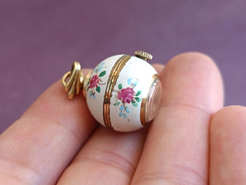 May include: A small, round, white pendant with a floral design in pink and blue. The pendant has gold accents and a gold-colored bail. The pendant is held in a hand.