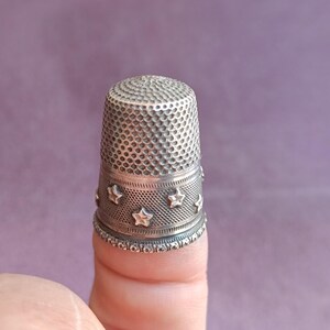 May include: A silver thimble with a textured top and a band of stars around the middle. The thimble is designed for sewing and crafting. The background is a solid purple color.