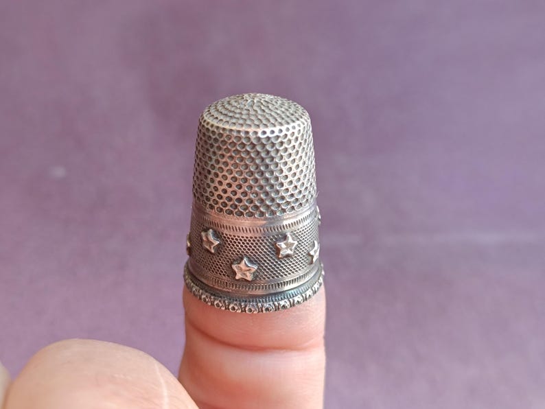 May include: A silver thimble with a textured top and a band of stars around the middle. The thimble is designed for sewing and crafting. The background is a solid purple color.