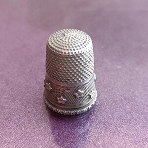 May include: A silver thimble with a textured surface and a band of star-shaped cutouts. The thimble is designed to protect the finger while sewing. The thimble is approximately 1 inch tall.