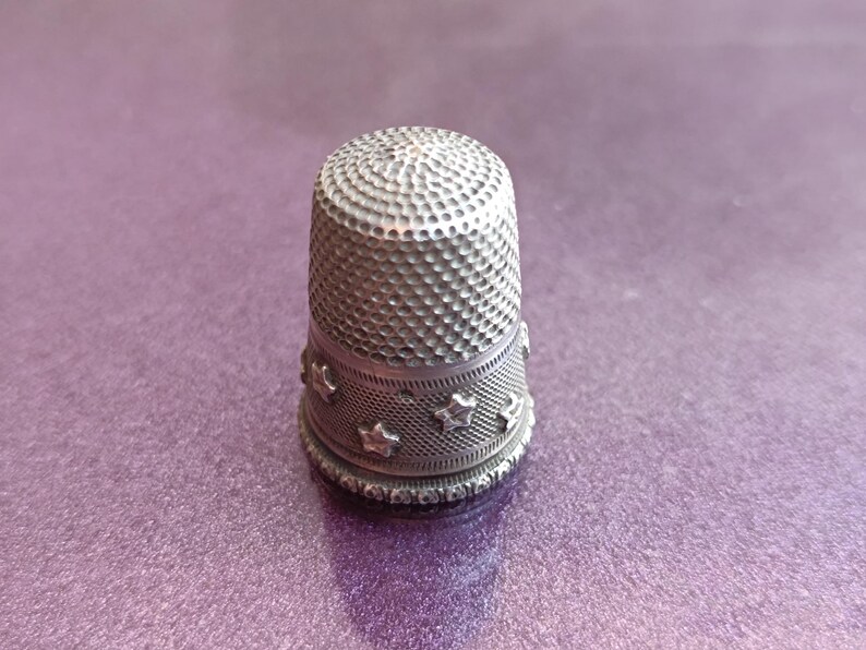 May include: A silver thimble with a textured surface and a band of star-shaped cutouts. The thimble is designed to protect the finger while sewing. The thimble is approximately 1 inch tall.
