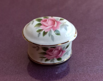 May include: A small, oval-shaped trinket box with a white background and a floral design. The box is decorated with pink roses and green leaves. The lid has a gold trim.