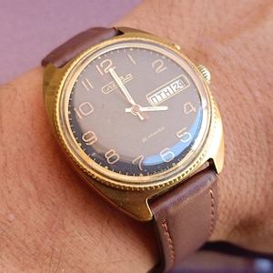Può includere: Un orologio da polso dorato con un cinturino in pelle marrone. Il quadrante dell'orologio è nero con numeri bianchi e una finestra della data che indica "ПТН 24". L'orologio è un orologio meccanico a 26 rubini.
