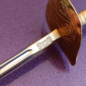 K&ouml;nnte beinhalten: Ein silbernes Schwert mit einem goldfarbenen Griff und einer Parierstange. Die Parierstange hat ein kompliziertes Design und die Klinge ist mit "WILKINSON SWORD" graviert.